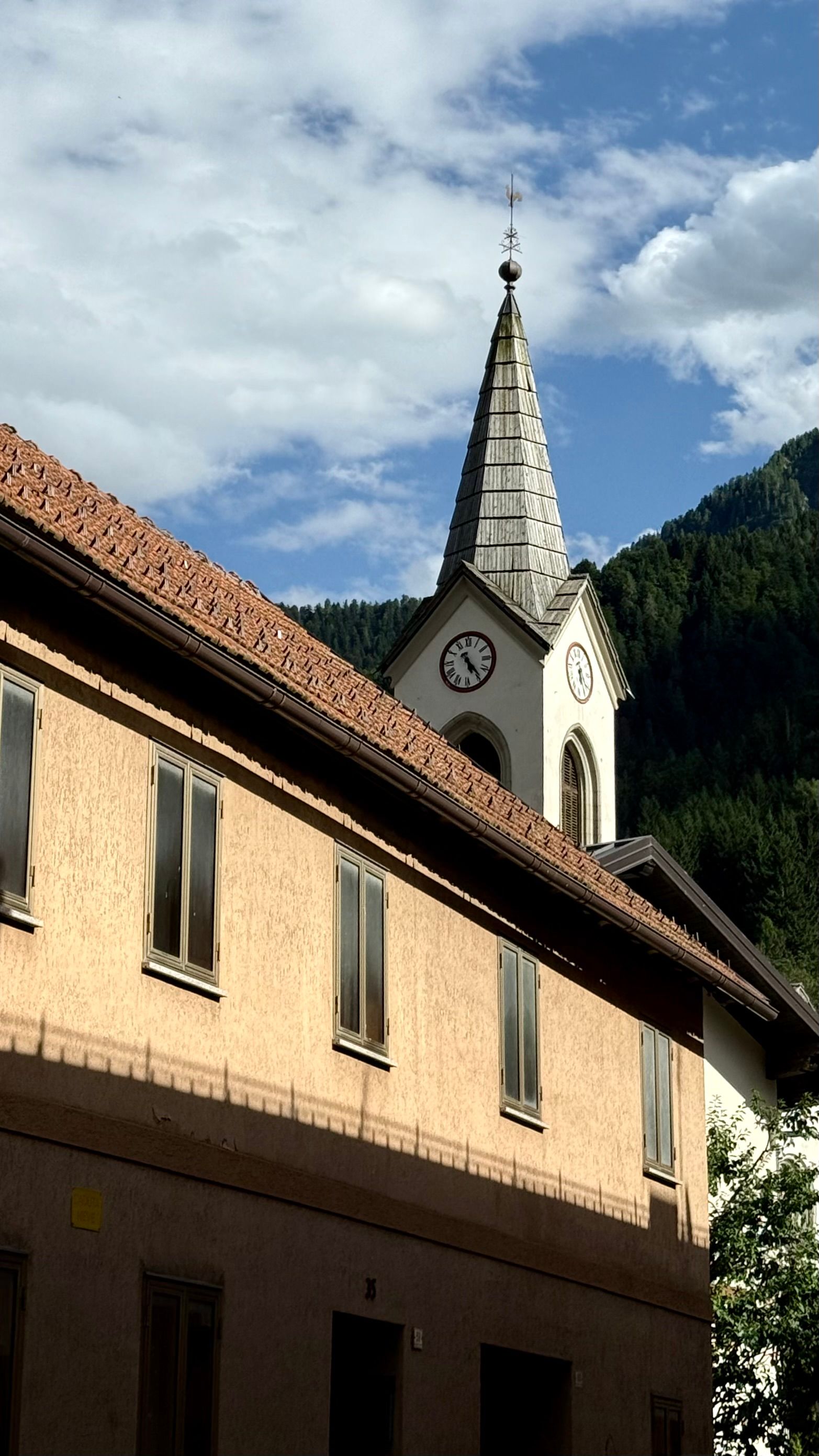 A picturesque Alpine church spire and building against a mountain backdrop in summer.