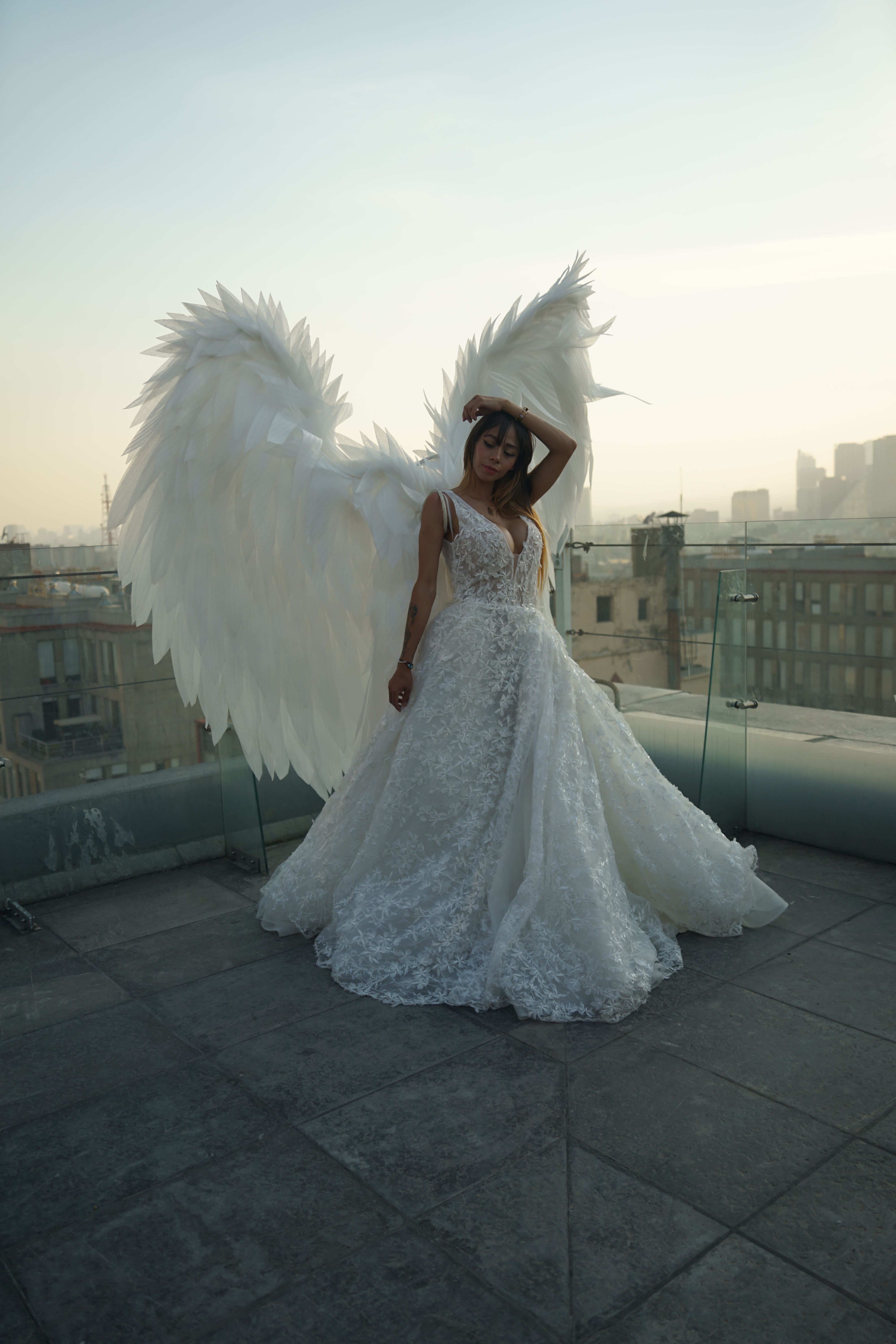 A woman in a white wedding dress with large angel wings stands on a rooftop against a city skyline at dusk.