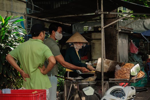 Street food vendor frying snacks outdoors in a bustling Asian market, interacting with customers.