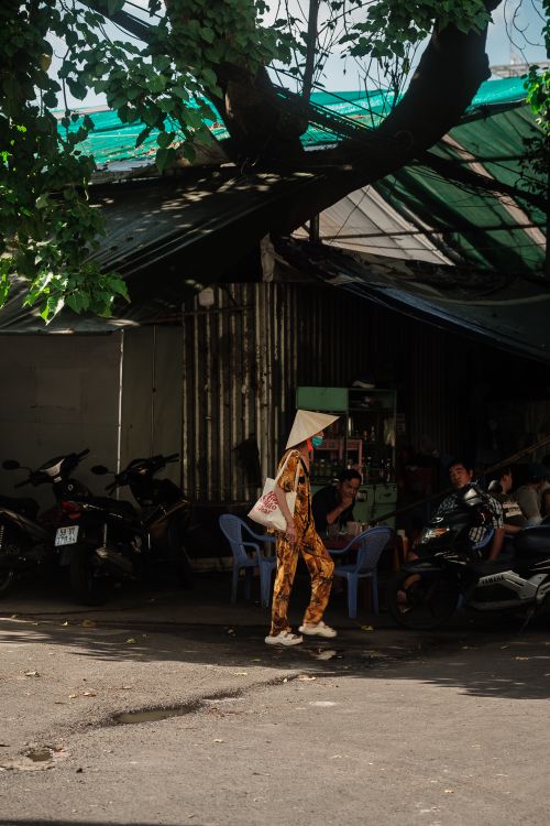 Free A woman in a conical hat walks past a group of men and mopeds under a tree shade. Stock Photo