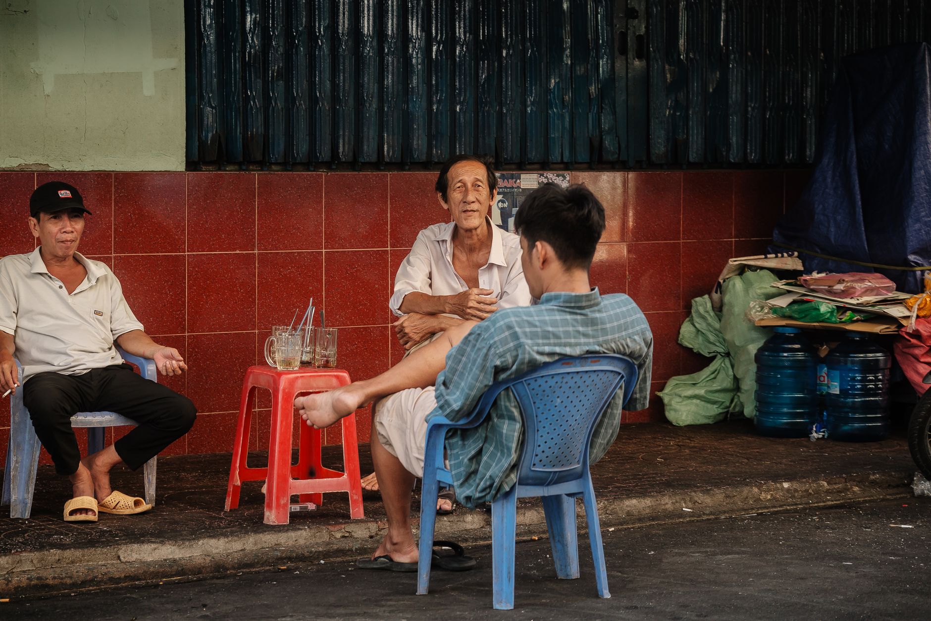 Three men sitting and chatting outdoors on red stools by the street.