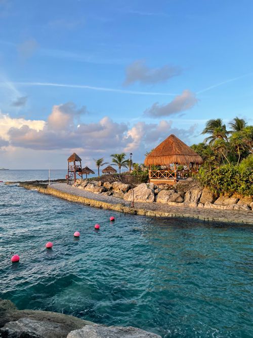 Free Idyllic tropical seaside scene featuring thatched huts, clear blue water, and a vibrant sunset sky. Stock Photo