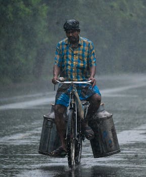 A man riding a bicycle in heavy rain while transporting large milk containers on a rural road.