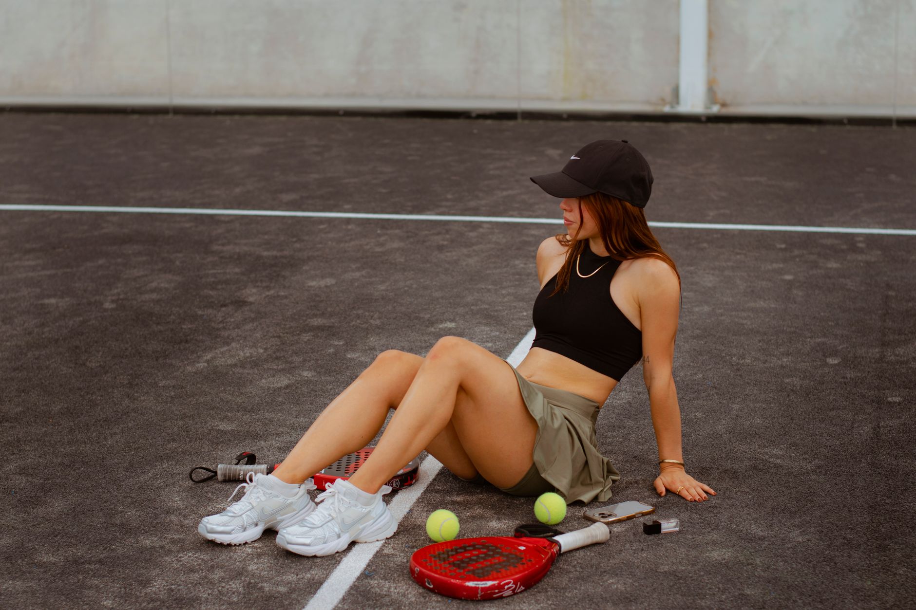 Young woman sitting on a tennis court with rackets and tennis balls, enjoying a relaxed moment outdoors.