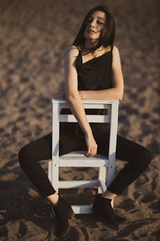 一位身着黑色服装的时尚女子坐在沙滩的白色椅子上，气质轻松