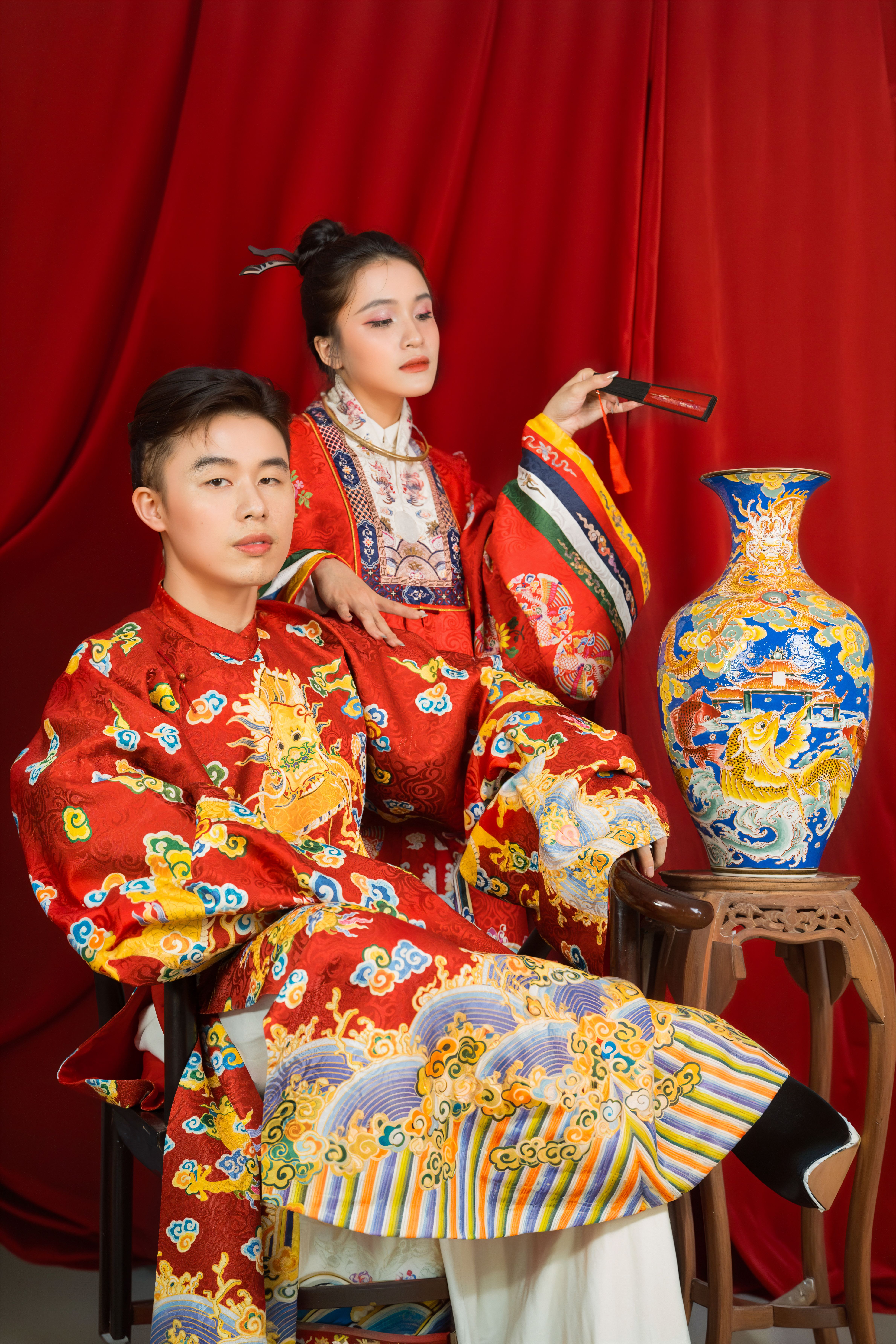Portrait of two adults in traditional Asian outfits beside a vividly painted vase.