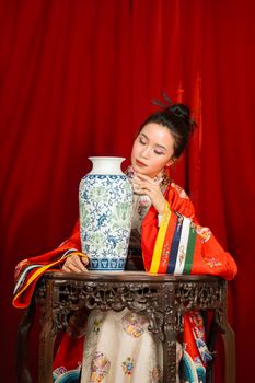 Woman in traditional dress sitting with ornate vase against red backdrop.
