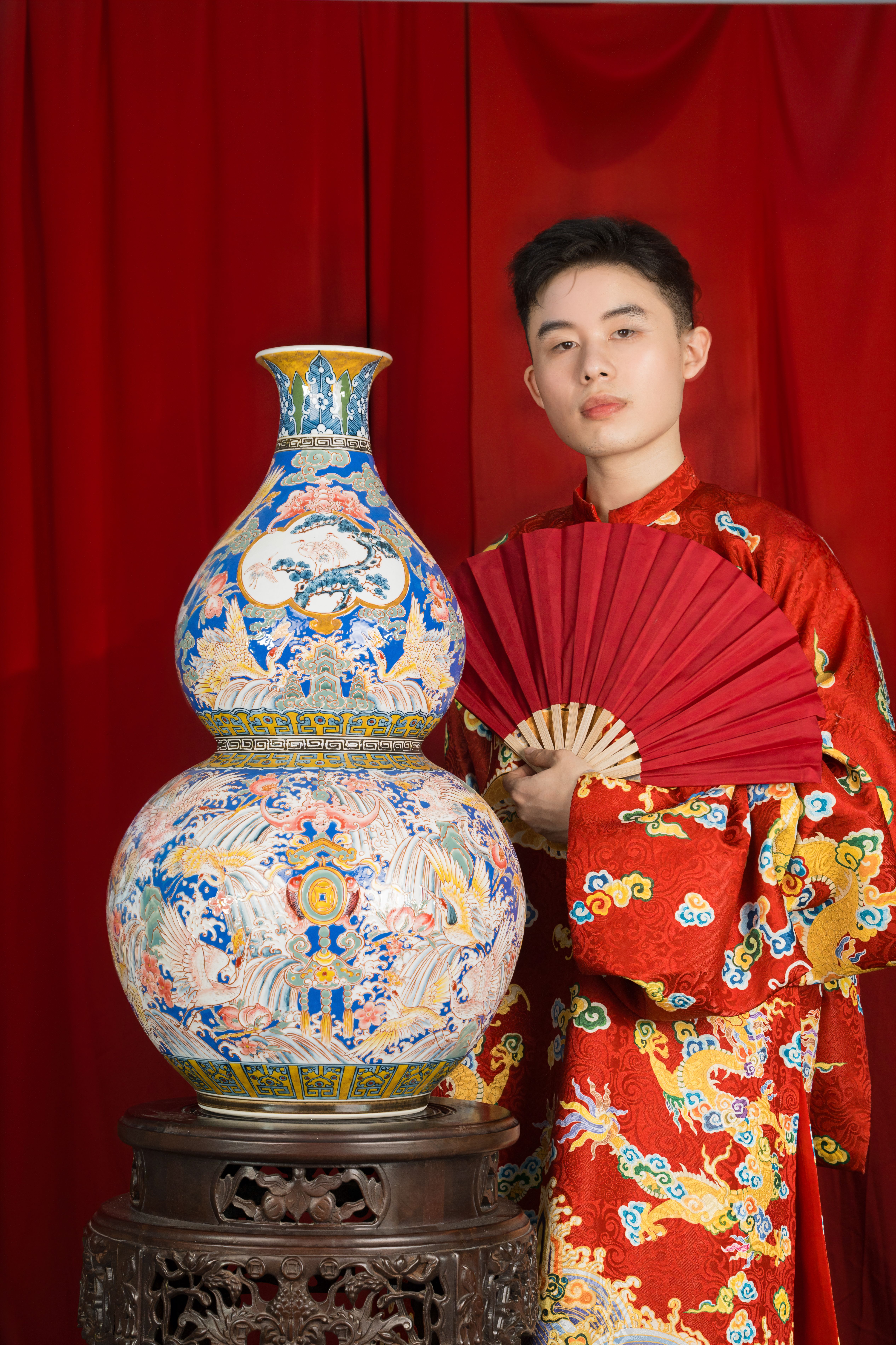 Asian man in traditional attire posing with a red fan and ornate vase against a red backdrop.