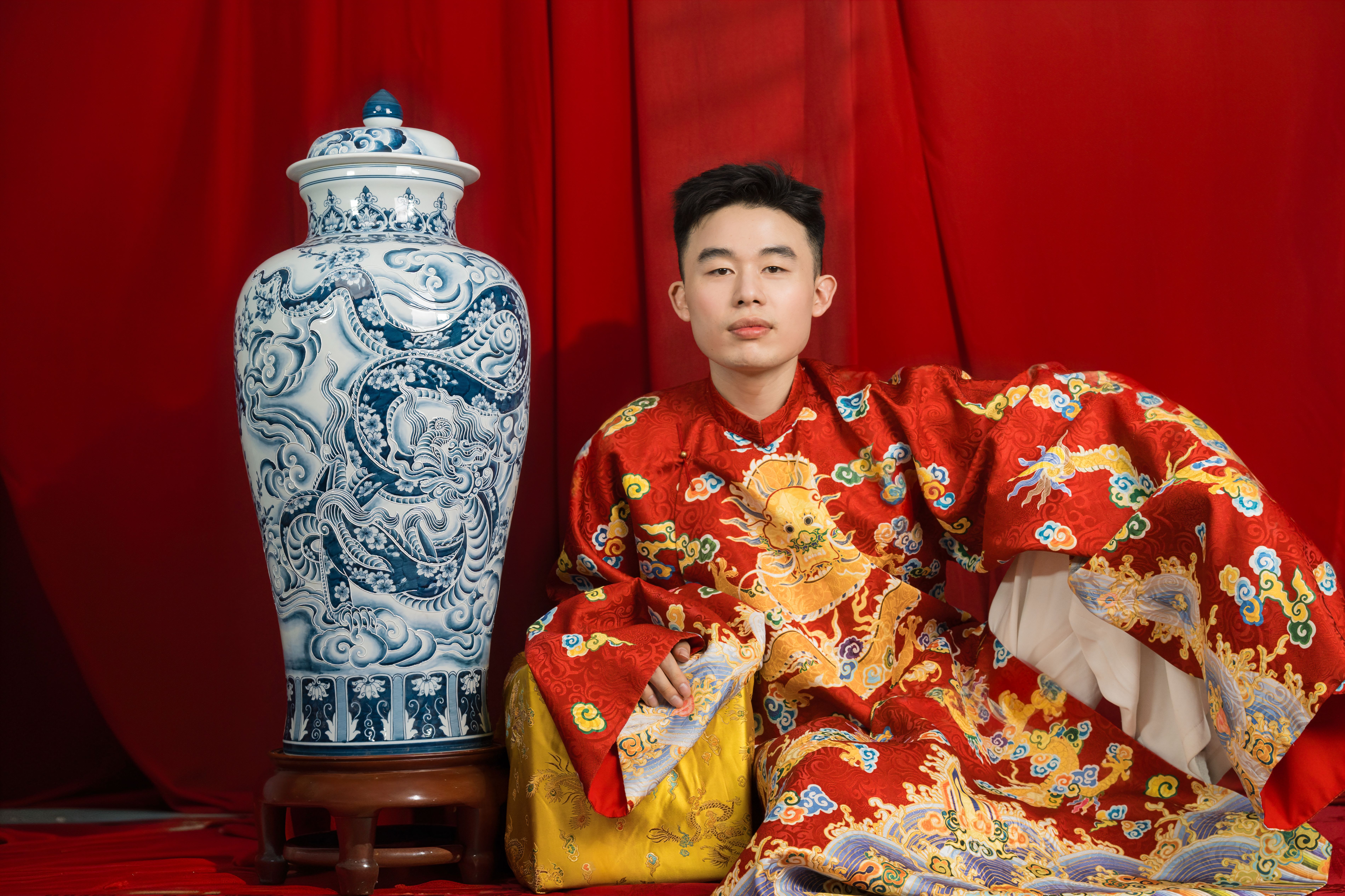 A young man in ornate Asian traditional clothing beside a blue and white ceramic vase with a red backdrop.