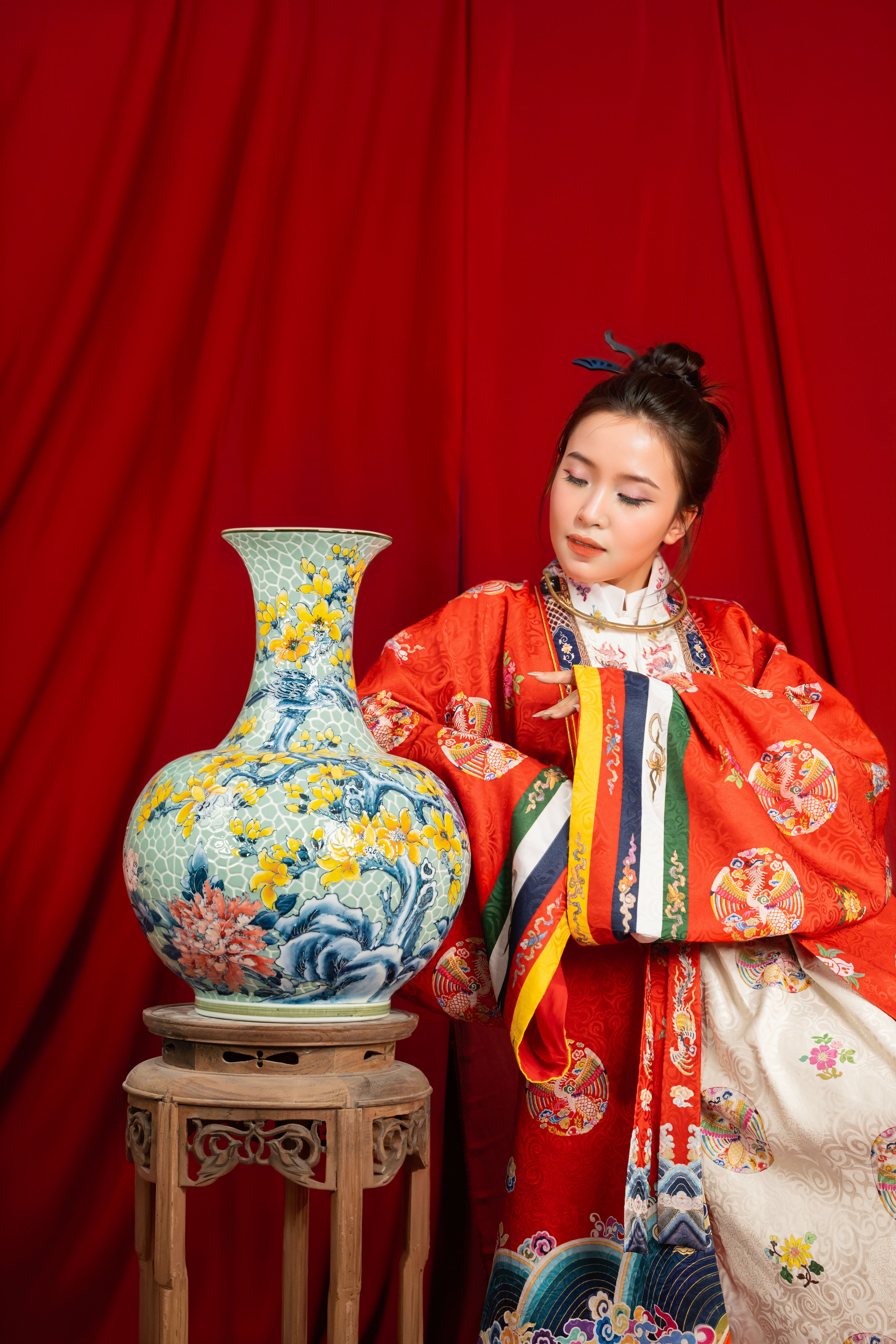 A woman in traditional attire poses beside an ornate ceramic vase on a wooden stand, set against a rich red backdrop.