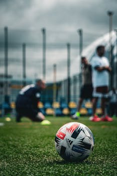 Focused soccer ball on grass field with players in the background during a cloudy day in Kyiv.