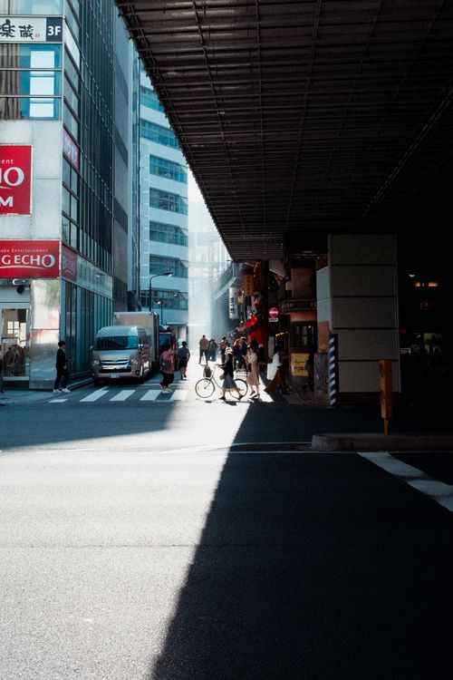 Free Vibrant Tokyo street with pedestrians and a cyclist under the cityscape's shadows. Stock Photo