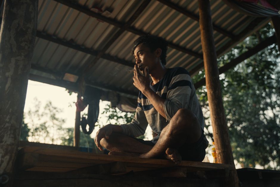 A young man in casual attire smokes while sitting on a rustic porch, reflecting in the soft evening light.