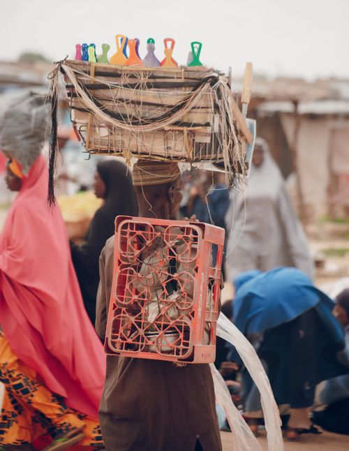Free A street vendor carrying goods on their head and back at a lively outdoor market, showcasing local culture. Stock Photo