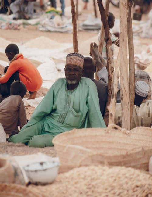 Free Man in traditional attire seated at a bustling outdoor market, surrounded by goods and people. Stock Photo