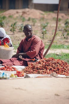 African vendor sitting by fresh produce outdoors, possibly selling vegetables in a local market.