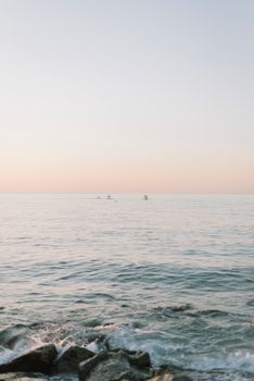Calm ocean at sunset with distant paddleboarders, rocks in foreground.