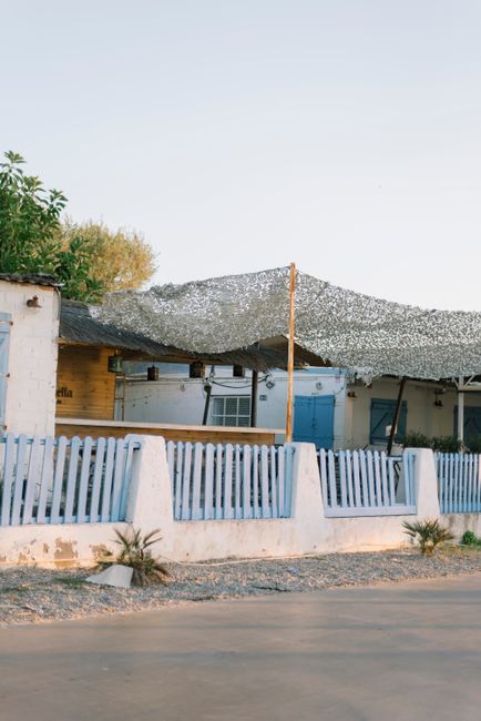 Rustic seaside cottage with blue fence and shaded patio, warm afternoon light.