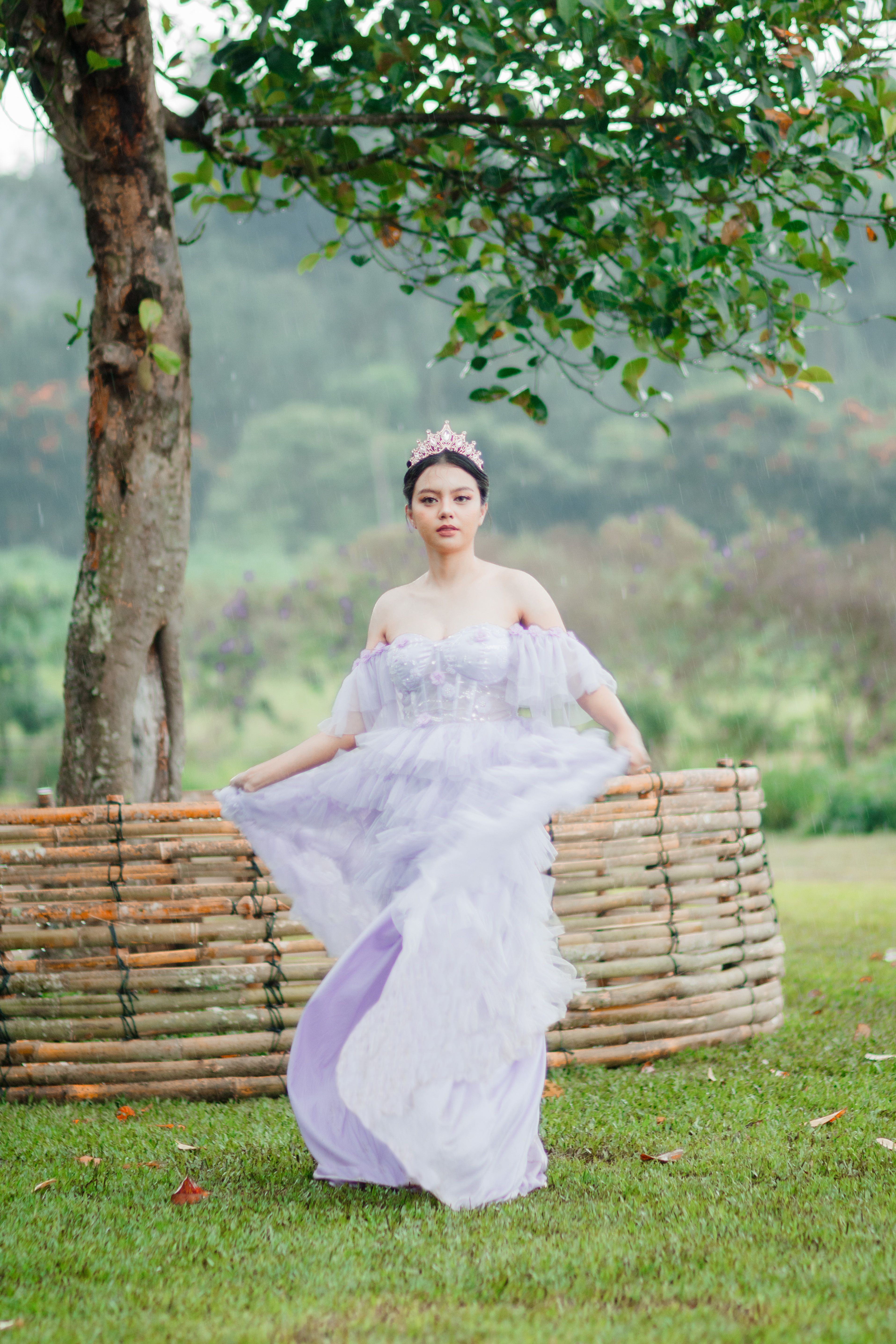 Graceful lady in lavender dress poses outdoors under a tree, evoking elegance.