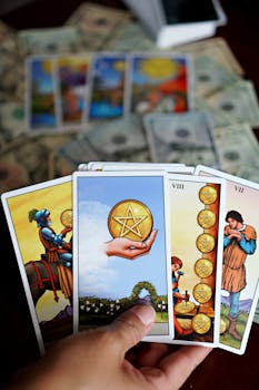 A hand holding tarot cards with money and tarot cards spread on a table.