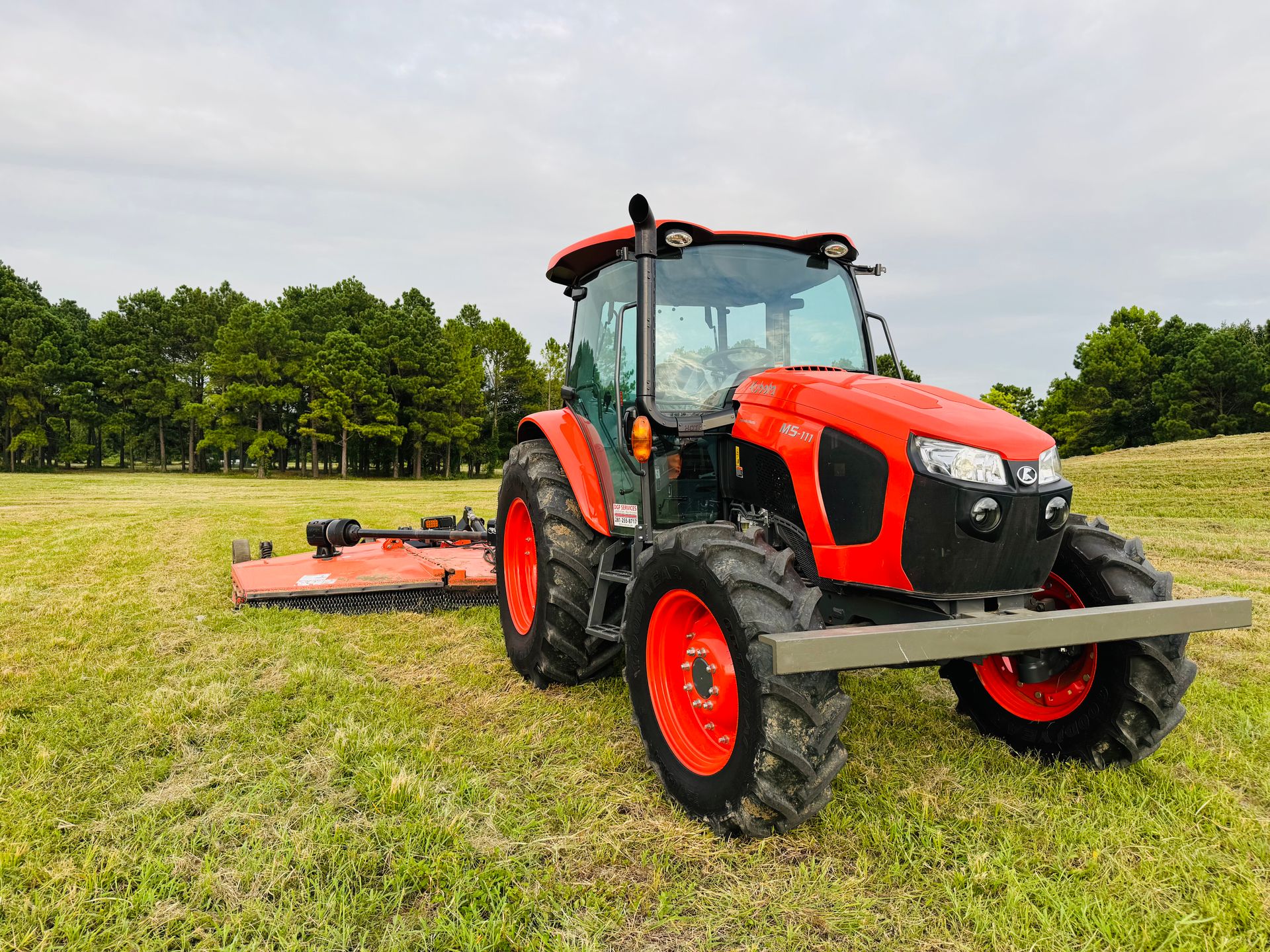 A red tractor with attached mower in a green field with trees in the background.