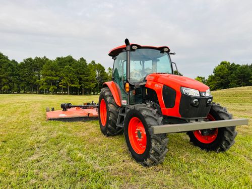 Free A red tractor with attached mower in a green field with trees in the background. Stock Photo
