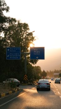 Cars on highway towards Muğla at sunset with road signs and trees silhouetted against golden light.