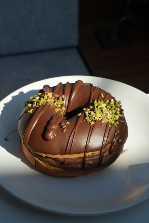 Free Close-up of a chocolate-covered donut topped with chopped pistachios on a white plate. Stock Photo