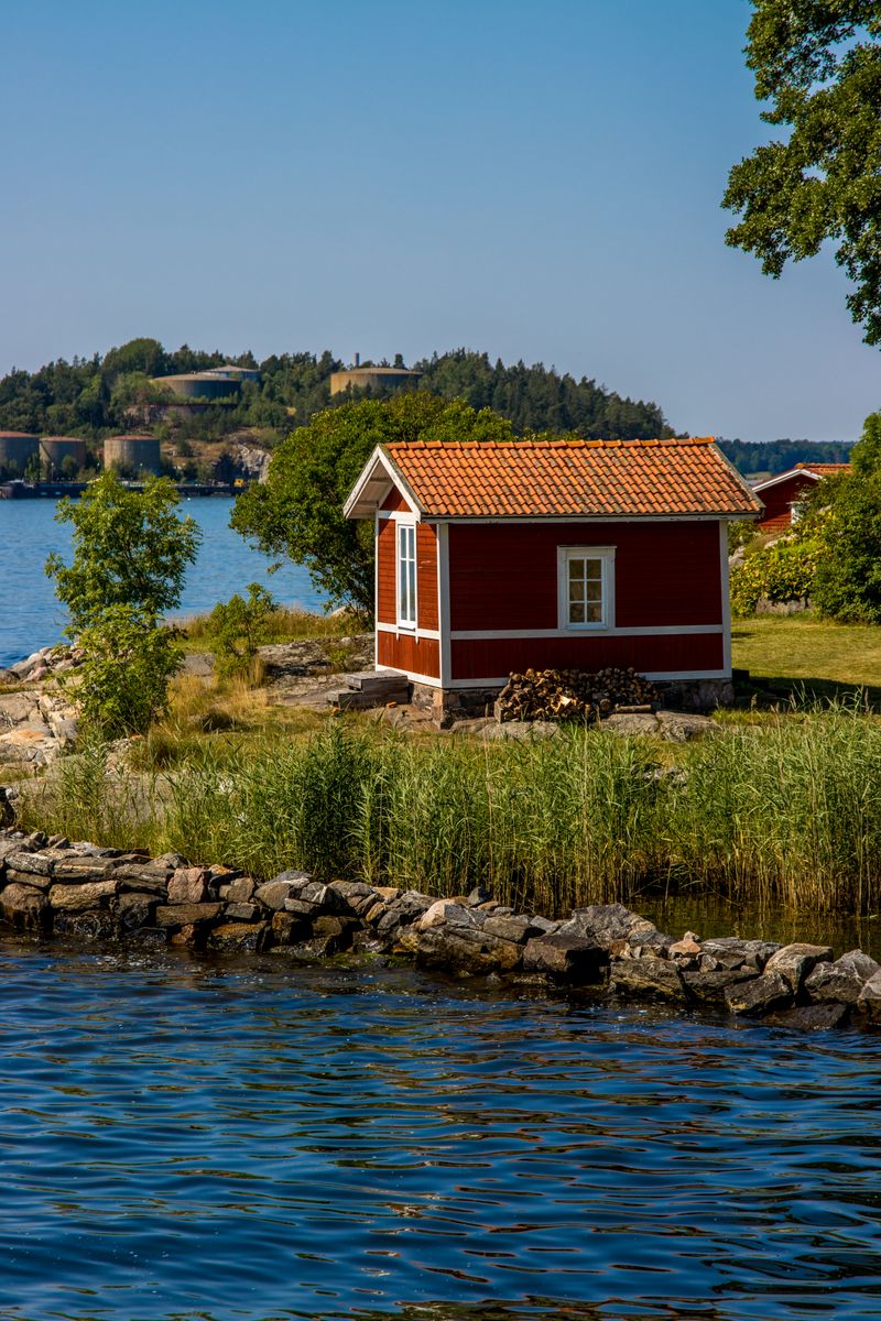 A small red cabin with an orange tiled roof on a green island with a stone shore