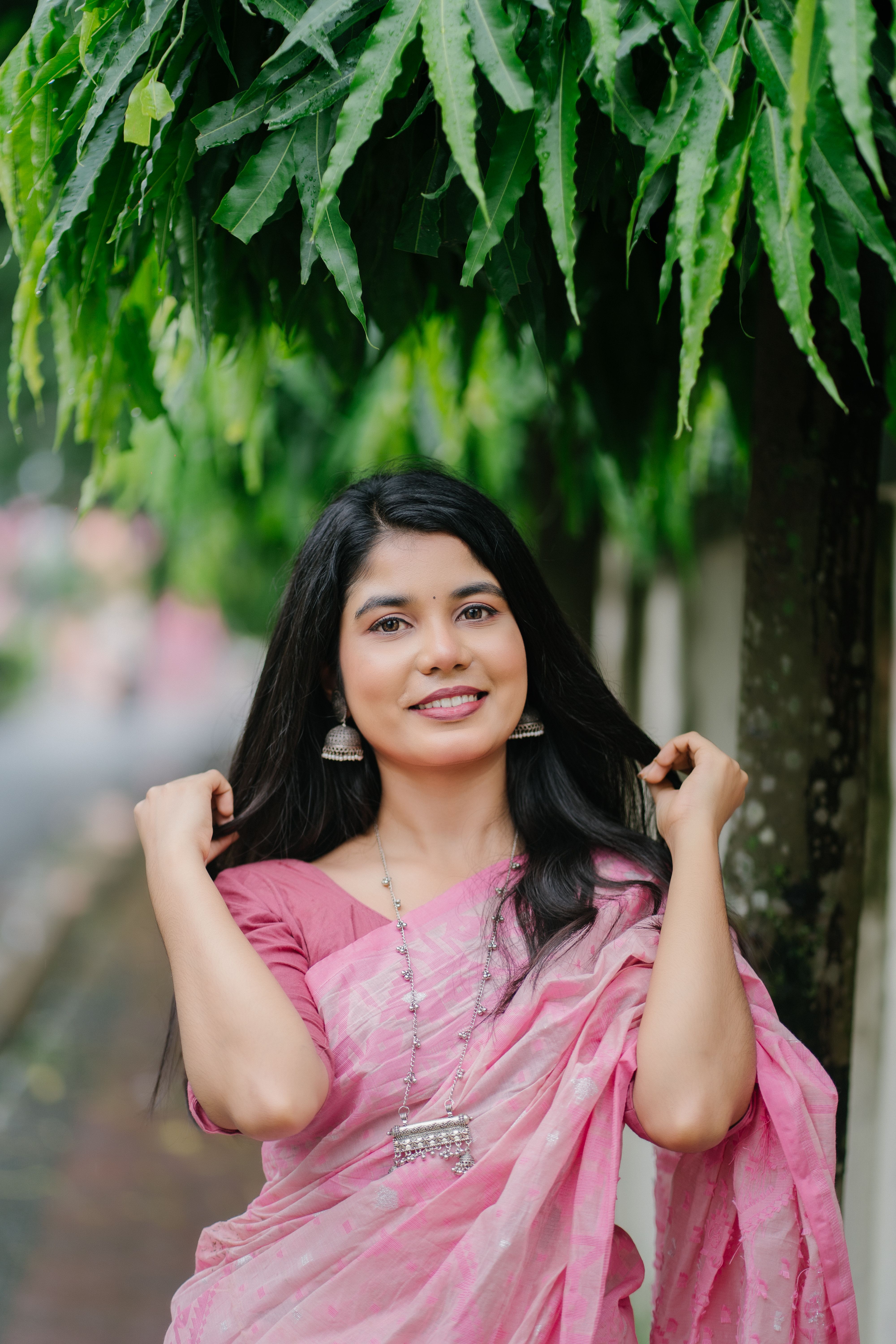 Smiling woman wearing a pink saree and jewelry under leafy trees.