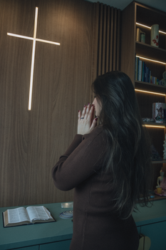A woman prays in a serene room with an illuminated cross and open Bible.
