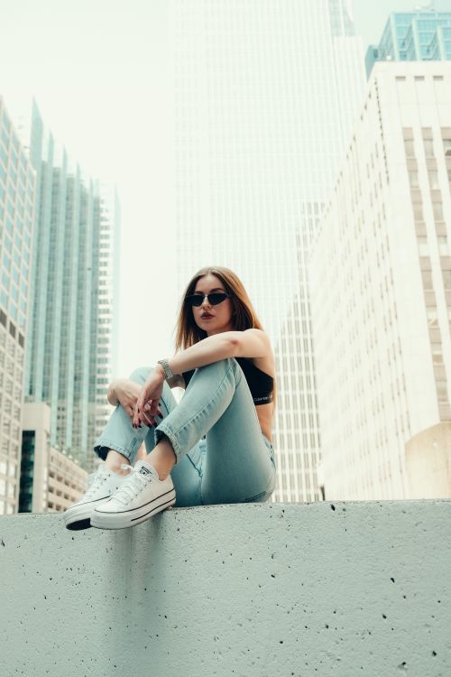 Free A stylish young woman in sunglasses sits on a rooftop with city skyscrapers in the background. Stock Photo