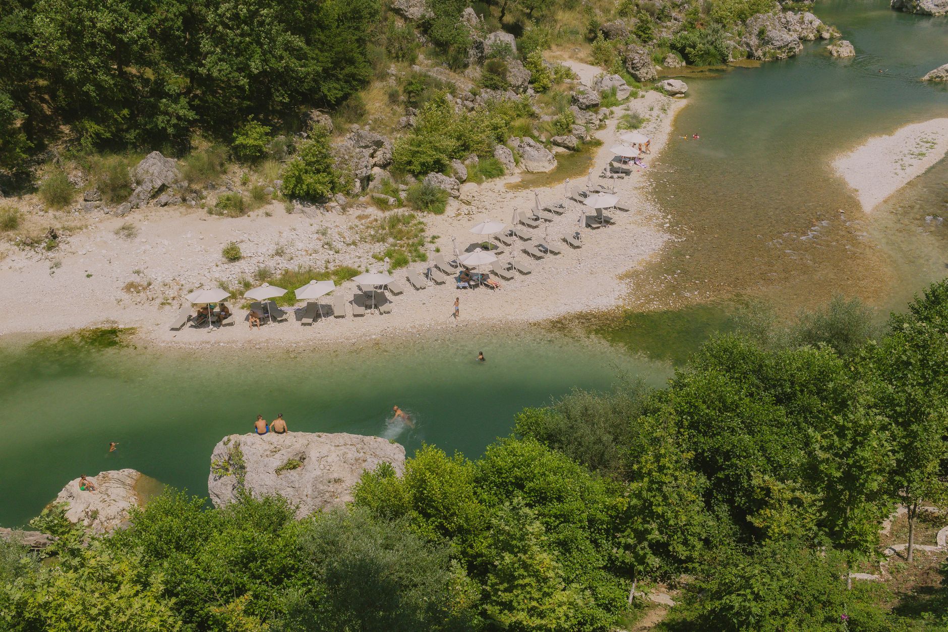 Peaceful riverside scene with sun umbrellas and lush greenery in Tepelenë, Albania.