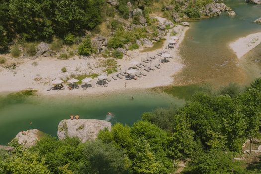 Peaceful riverside scene with sun umbrellas and lush greenery in Tepelenë, Albania.