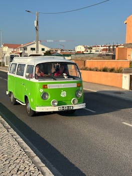 A classic green Volkswagen camper van driving down a sunny street in a suburban neighborhood.