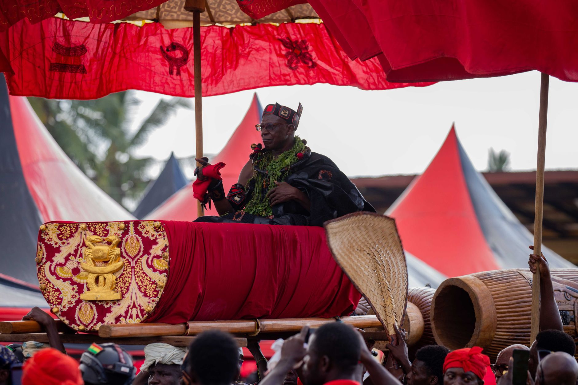A king in ceremonial attire sits under a vibrant red canopy during a traditional African festivity.