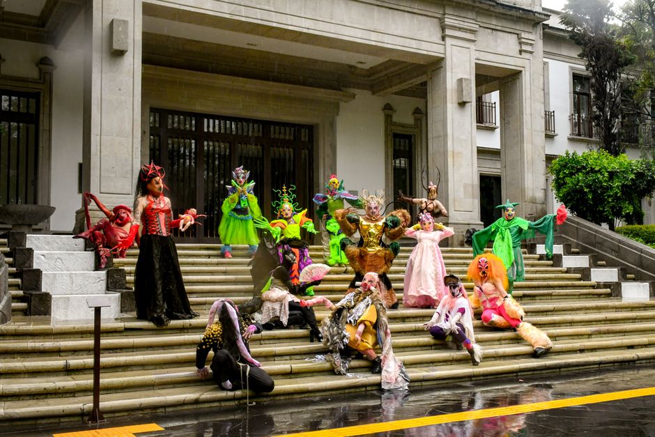 Vibrant costumed performers pose on majestic mansion steps, showcasing elaborate outfits.