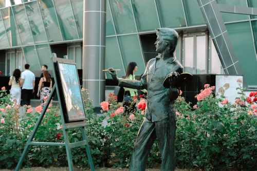 Free A bronze artist statue amidst rose bushes with people nearby outside a modern building. Stock Photo