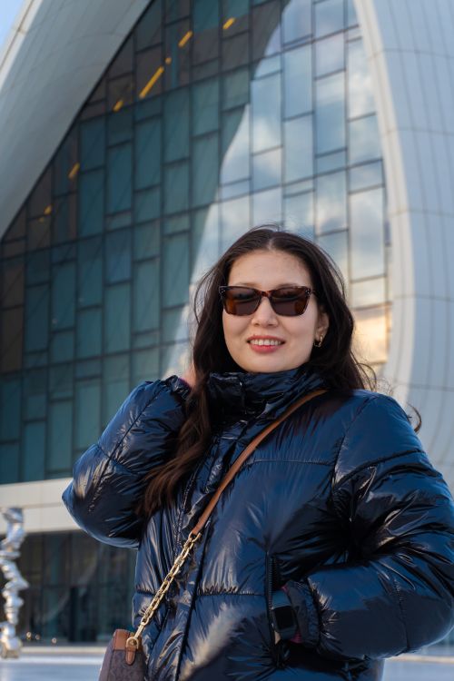 Free Woman smiling in puffer jacket and sunglasses in front of modern building. Urban fashion scene. Stock Photo