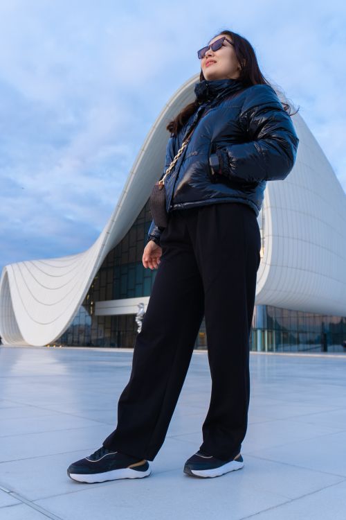Free A confident woman in winter attire poses before a stunning modern building at dusk. Stock Photo