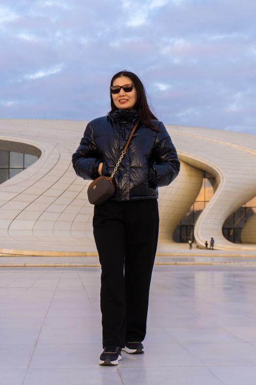 Free Woman in sunglasses and black jacket walking by modern architectural building at sunset. Stock Photo