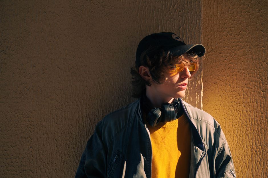 Teenager in stylish outfit and headphones standing against a textured wall in sunlight.