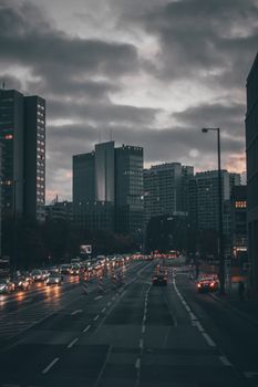 Stream of moving cars with head lights illuminating road of contemporary city past multistory buildings in overcast evening