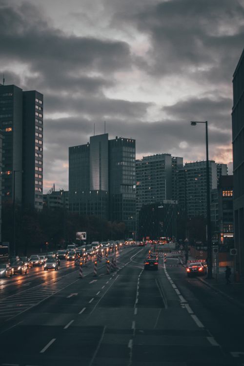 Free Stream of moving cars with head lights illuminating road of contemporary city past multistory buildings in overcast evening Stock Photo