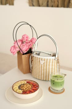 Chic breakfast setup with smoothie, handbag, and roses on a white table.