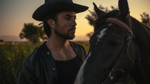 Free A cowboy in a hat stands beside his horse during a sunset in an open field. Stock Photo
