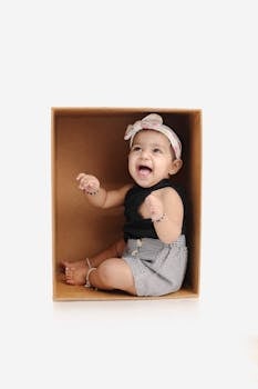 Cute baby playing inside a box, dressed in black top and plaid shorts, with a bow headband.
