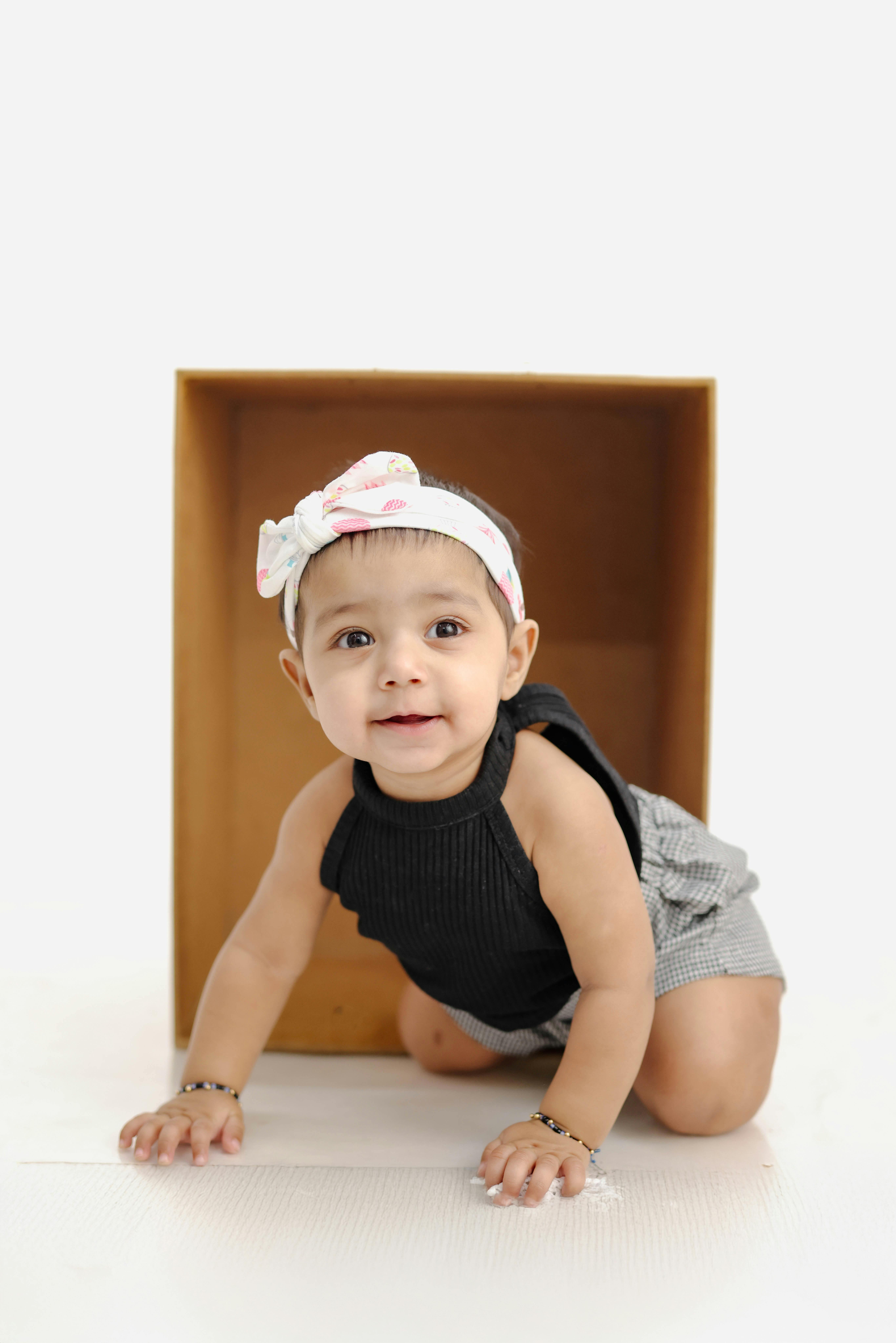 Cute baby girl wearing headband crawling out of wooden box against white background.