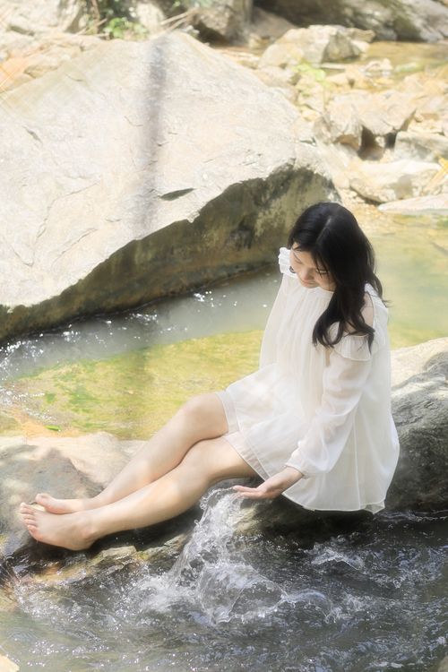 Free Serene woman in white dress sits by a gentle stream, enjoying tranquility. Stock Photo