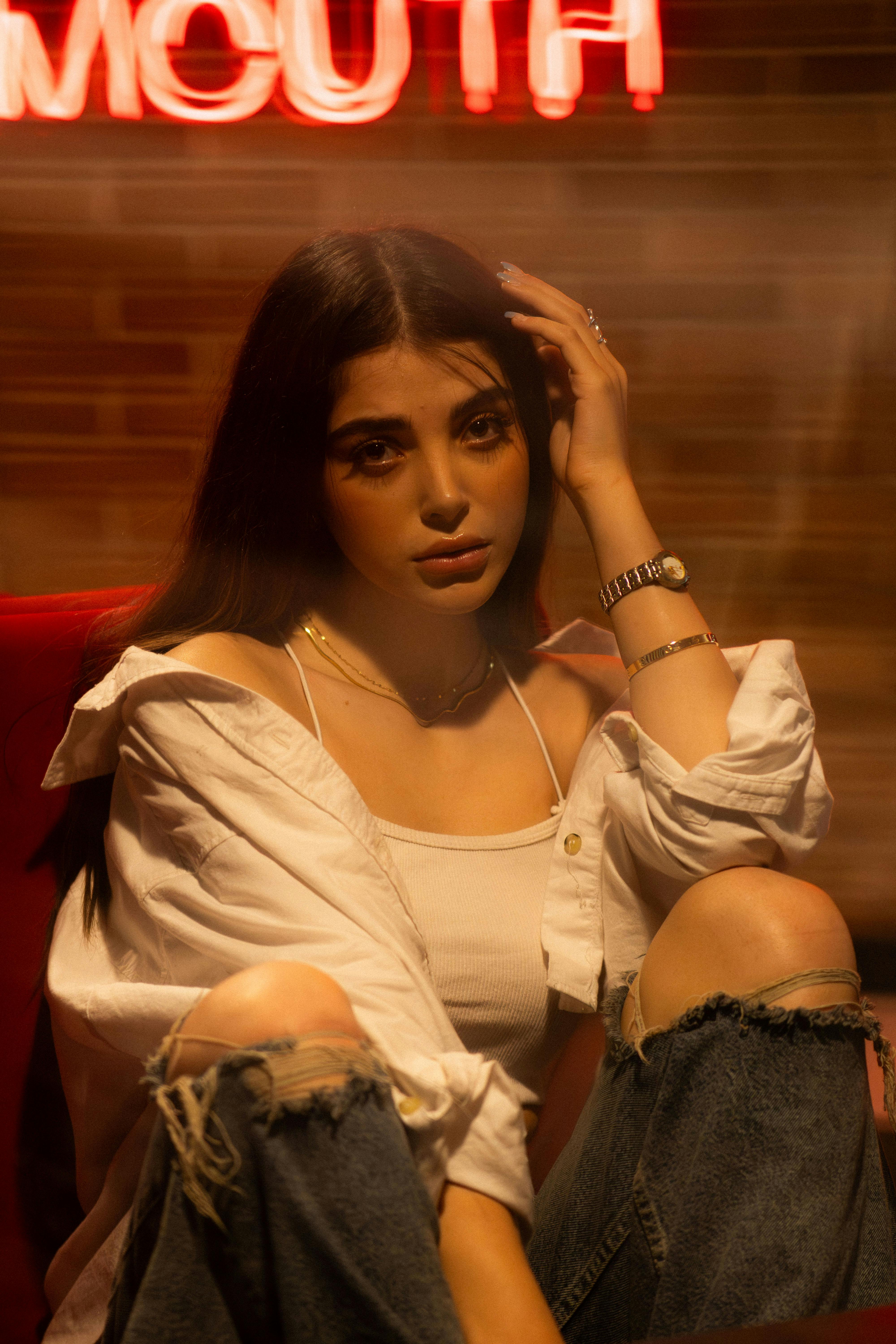 Stylish portrait of a young woman sitting indoors, wearing casual fashion in moody lighting.